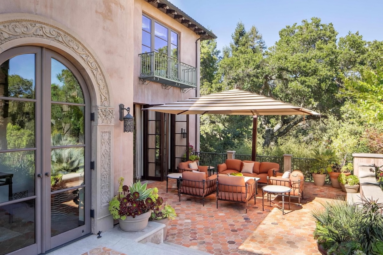 This elegant outdoor patio features a classic Mediterranean aesthetic with terracotta-toned hexagonal floor tiles and a warm, stucco-finished home exterior. The space is furnished with comfortable, rust-colored cushioned seating arranged under a large, neutral-toned cantilevered umbrella, creating an inviting area for relaxation. A wrought-iron balcony above and lush, mature trees in the background enhance the serene, private atmosphere of this outdoor living space.