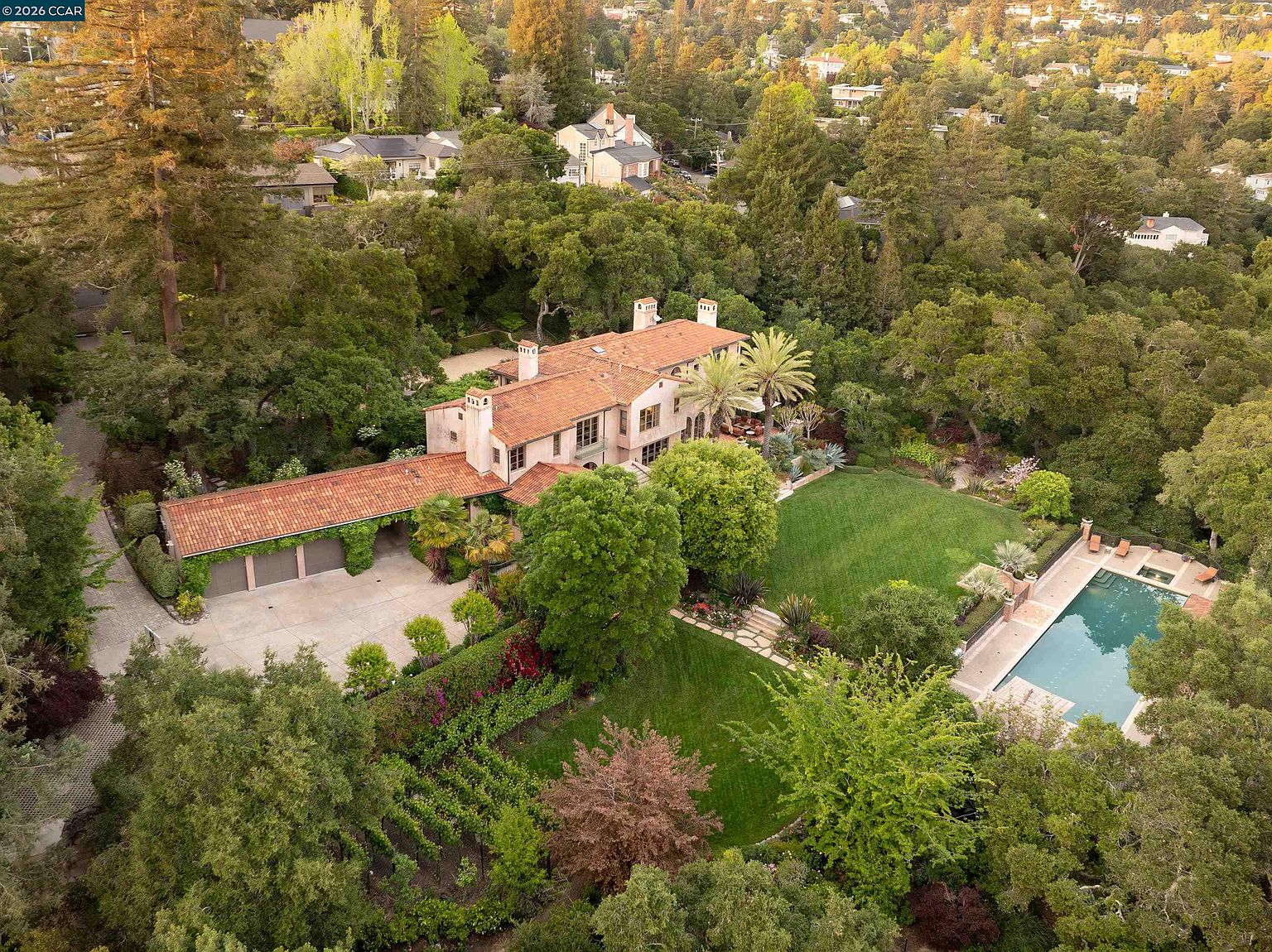 This high-angle aerial view showcases a sprawling Mediterranean-style estate nestled within a lush, wooded landscape. The property features a multi-level residence with a terracotta tile roof, a detached garage, a manicured lawn, and a private swimming pool. The surrounding mature trees and garden beds create a secluded, park-like atmosphere, emphasizing the home's expansive outdoor living spaces.
