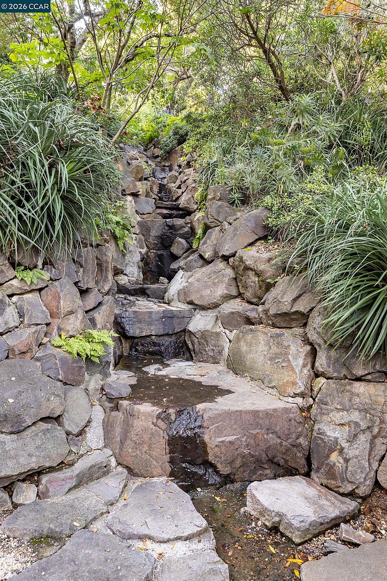 This image captures a tiered stone water feature integrated into a lush, naturalistic garden setting. Large, rugged boulders form a cascading staircase for the water, flanked by dense foliage, ferns, and ornamental grasses that create a serene, woodland atmosphere. The perspective is a low-angle, upward-looking shot that emphasizes the verticality and organic texture of the rockwork.