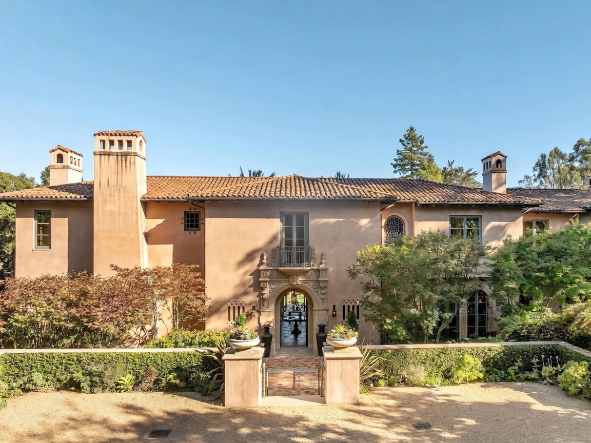 This Mediterranean-style estate features a warm, stucco-clad facade with a classic red-tiled roof and prominent chimneys. A grand, ornate stone archway serves as the primary entrance, leading to a courtyard visible through the open doorway, while a manicured hedge and stone pillars frame the foreground. The symmetrical design and lush landscaping create an elegant and inviting curb appeal.