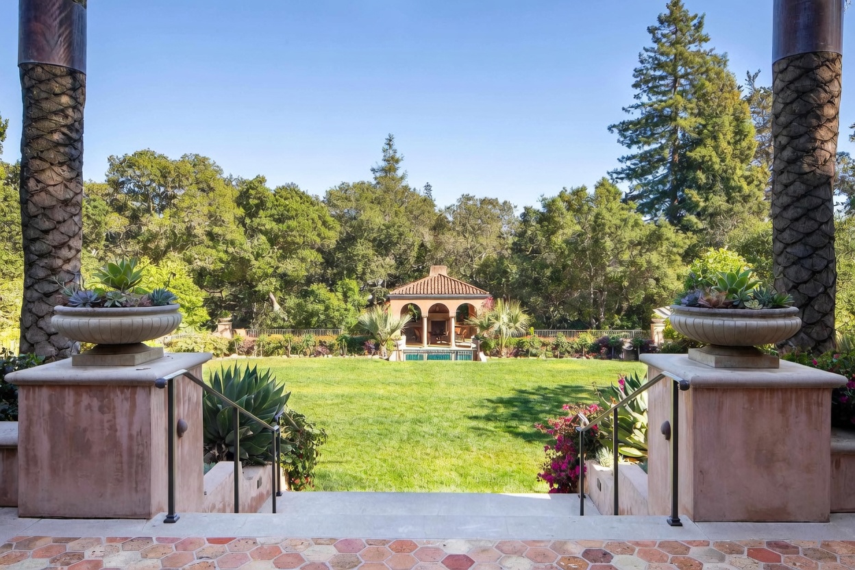 This perspective captures a beautifully landscaped backyard from an elevated stone patio, looking down a set of stairs toward a lush green lawn. The scene is framed by two large stone pillars topped with decorative planters filled with succulents, leading the eye toward a charming, open-air pavilion with a tiled roof at the far end of the garden. The surrounding mature trees and vibrant flowering bushes create a serene, private atmosphere perfect for outdoor relaxation.
