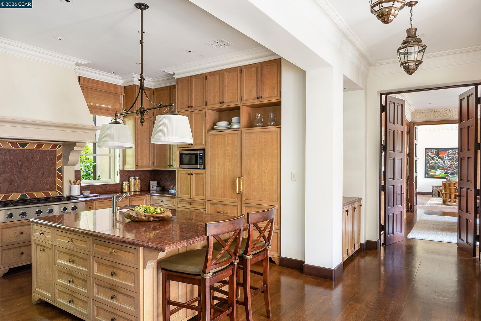 This elegant kitchen features light wood cabinetry, a large central island with a dark stone countertop, and a matching stone backsplash behind the range. The space is illuminated by dual pendant lights hanging over the island and recessed ceiling lighting, creating a warm and inviting atmosphere. A wide doorway leads into an adjacent room, showcasing a view of a hallway and a framed piece of art, while the overall design blends traditional warmth with functional luxury.