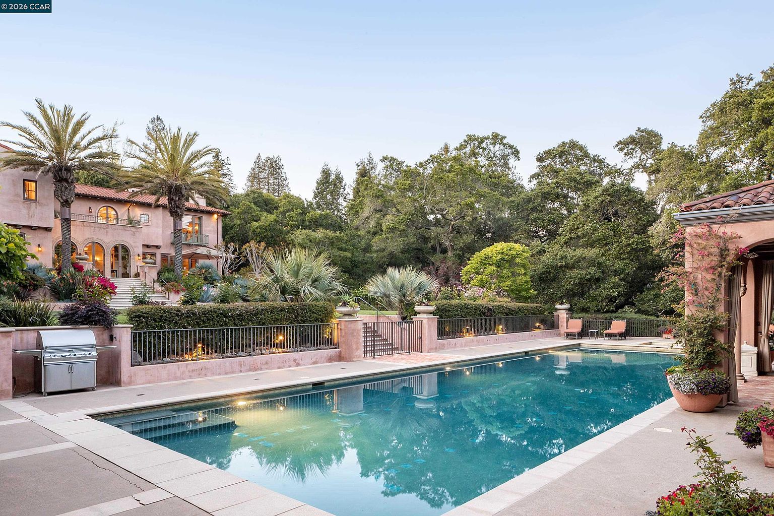 This expansive Mediterranean-style estate features a large, rectangular swimming pool as the centerpiece of the backyard. The property includes a multi-level landscape with lush greenery, palm trees, and a grand stucco residence in the background with arched windows and a tiled roof. A stainless steel grill is positioned on the patio, and the scene is framed by a wrought-iron fence, creating a serene and luxurious outdoor living environment.