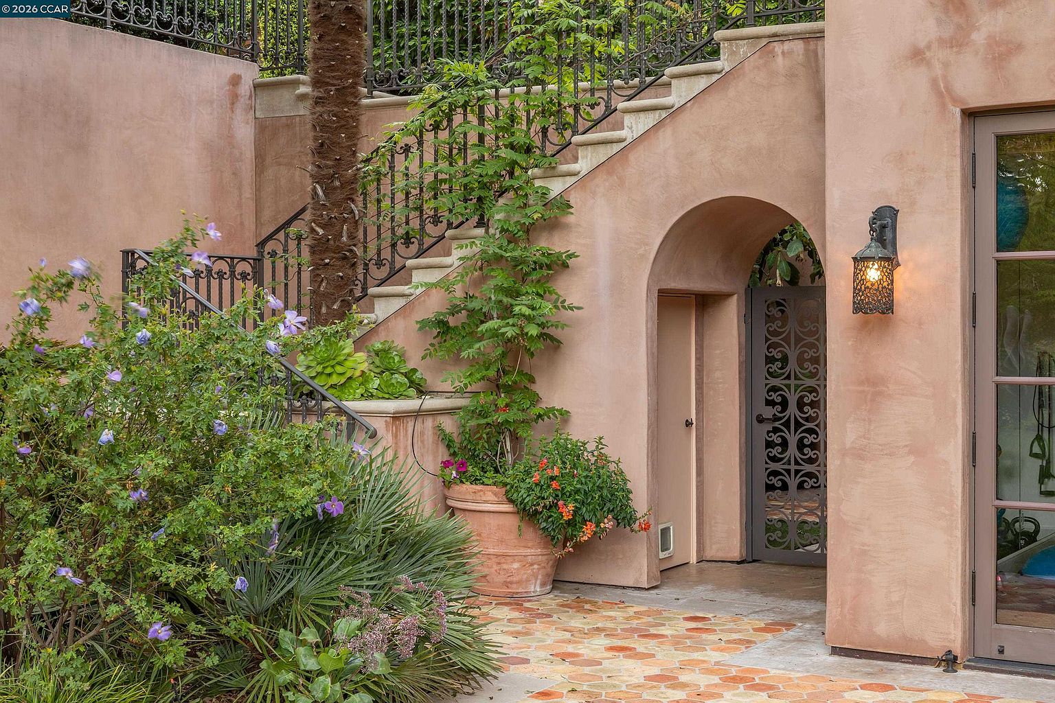 This charming Mediterranean-style garden area features a terracotta-tiled patio leading to an arched entryway with an ornate metal gate. A stone staircase with intricate wrought-iron railings ascends along the side of the stucco building, accented by lush greenery, a large potted plant, and a flowering shrub. The warm, earthy tones and architectural details create an inviting and elegant outdoor atmosphere.