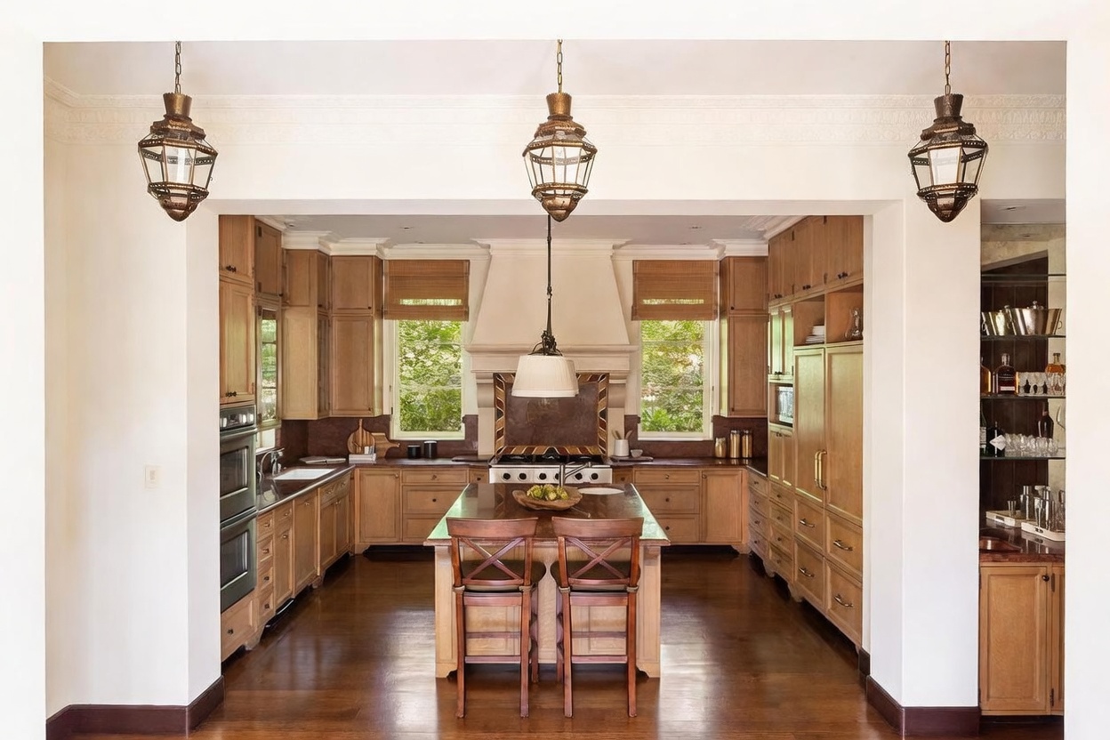This spacious, traditional-style kitchen features warm-toned wood cabinetry, a central island with bar seating, and a prominent range hood centered between two windows. The room is illuminated by three decorative hanging lanterns and recessed lighting, creating a welcoming and functional atmosphere. The perspective is a straight-on, symmetrical shot looking through a wide doorway into the heart of the kitchen.