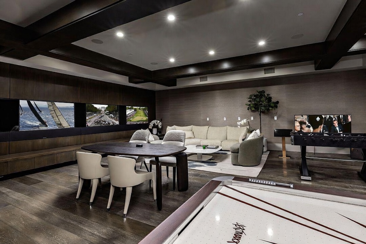This expansive, modern entertainment room features a sophisticated dark wood aesthetic with recessed lighting and coffered ceiling details. The space is furnished with a central dining table, a plush sectional sofa, a foosball table, and an air hockey table, creating a versatile recreational area. A wall of integrated screens provides a cinematic focal point, while the open layout and neutral color palette offer a luxurious and inviting atmosphere.