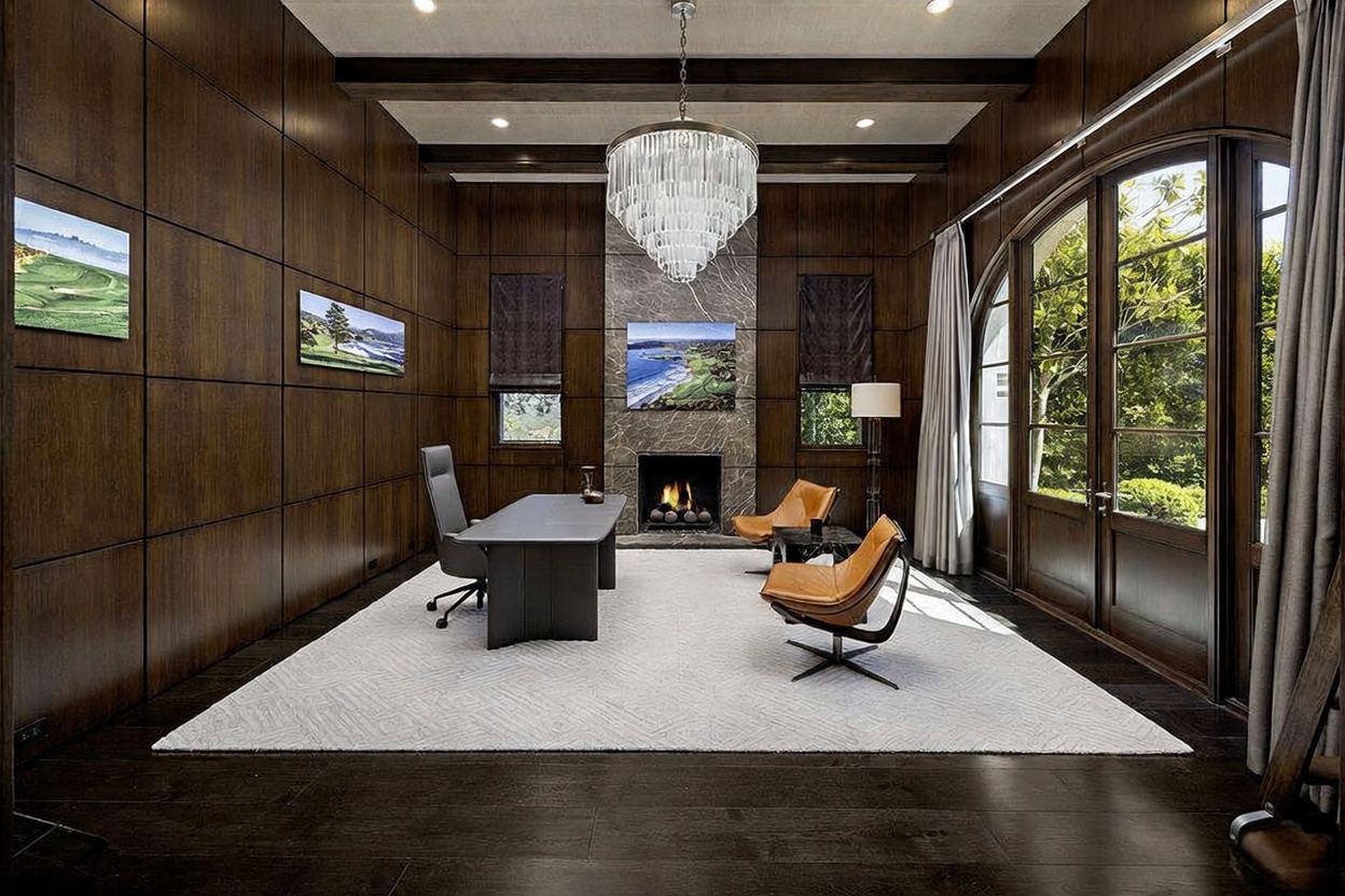This sophisticated home office features dark wood-paneled walls and a large, light-colored area rug centered in the room. A modern desk and two leather lounge chairs are arranged around a stone-clad chimney breast with a built-in fireplace. Tall, arched glass doors provide ample natural light and a view of the outdoors, while a grand crystal chandelier serves as a focal point above the workspace.