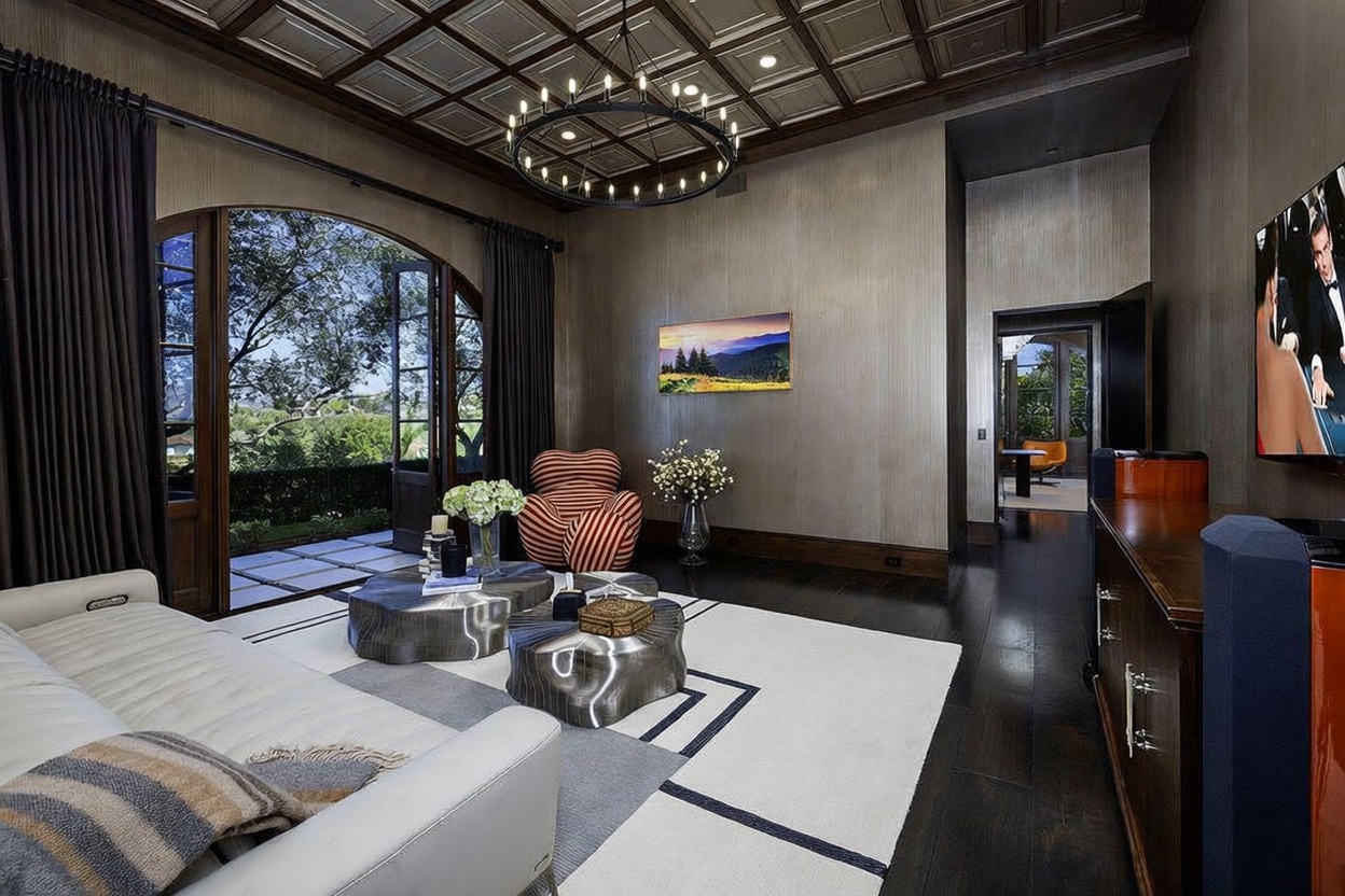 This sophisticated living room features a coffered ceiling, dark wood flooring, and a large arched doorway that opens to an outdoor patio. The space is furnished with a white sofa, a unique striped accent chair, and two metallic, organic-shaped coffee tables centered on a geometric area rug. A large television is mounted on the wall, and the room is accented by a modern circular chandelier and a view into an adjacent room.
