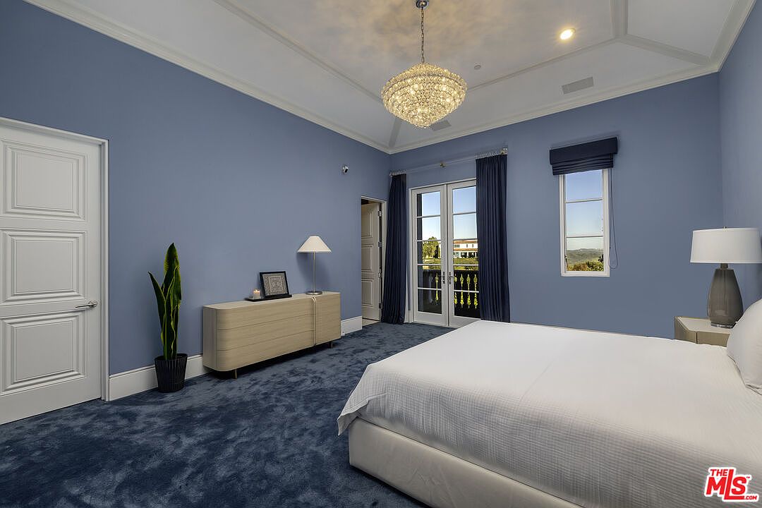 This spacious primary bedroom features soft blue walls, plush dark blue carpeting, and a high tray ceiling accented by a crystal chandelier. A large bed sits in the foreground, while a light wood dresser with a minimalist lamp and a tall potted plant occupy the corner. French doors with dark curtains open to a balcony, providing natural light and a view of the surrounding landscape.