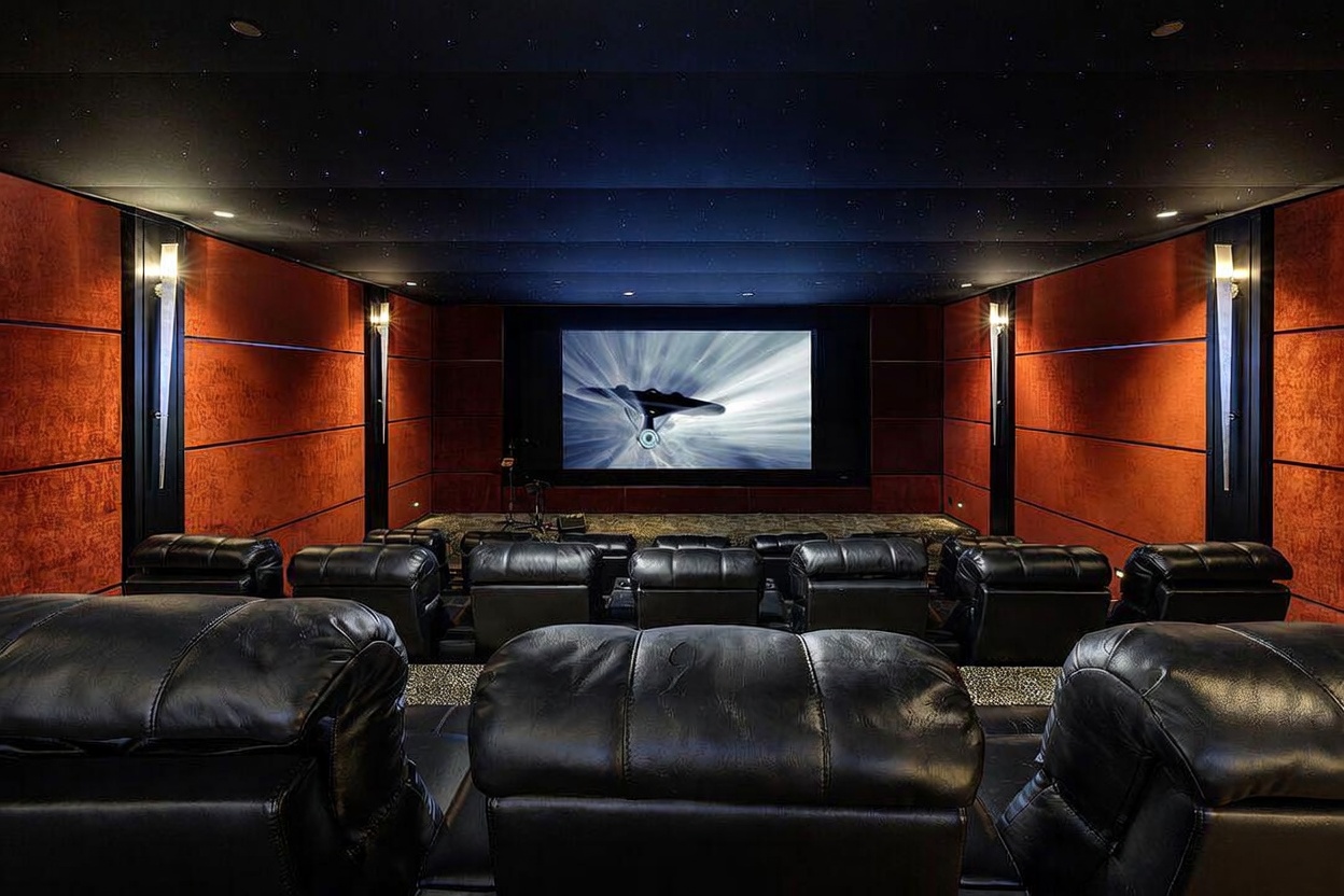 This dedicated home theater features multiple rows of plush, black leather reclining seats arranged for optimal viewing of a large projection screen. The room is designed with dark, sound-absorbing wall panels in a warm, textured finish and a ceiling accented with fiber-optic star lighting. The perspective is from the back of the room, looking forward toward the screen, creating an immersive and professional cinematic atmosphere.