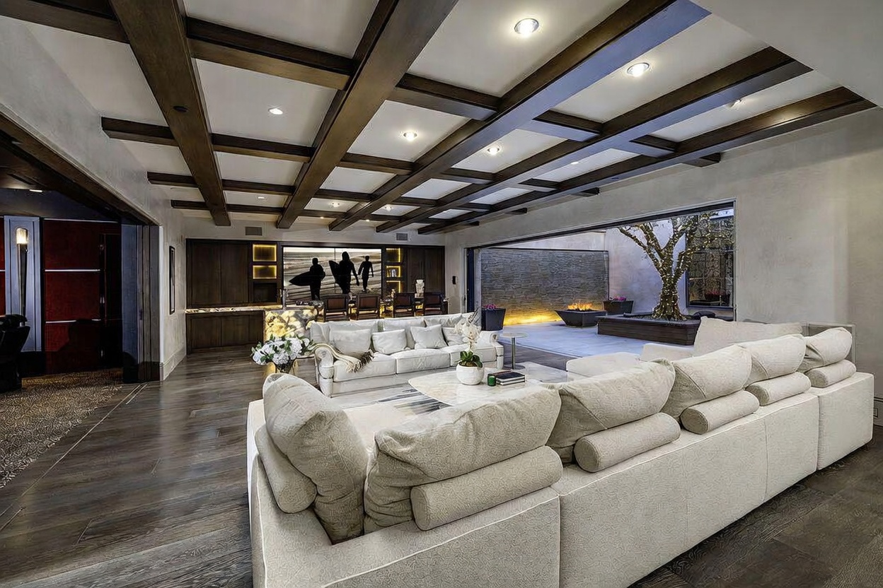This expansive, modern living room features a coffered ceiling with dark wood beams and recessed lighting, creating a sophisticated atmosphere. A large, light-colored sectional sofa dominates the foreground, while a wall of glass opens seamlessly to an outdoor patio area with a lit tree and a fire feature. The space also includes a bar area with a backlit counter and a large screen, blending luxury entertainment with indoor-outdoor living.