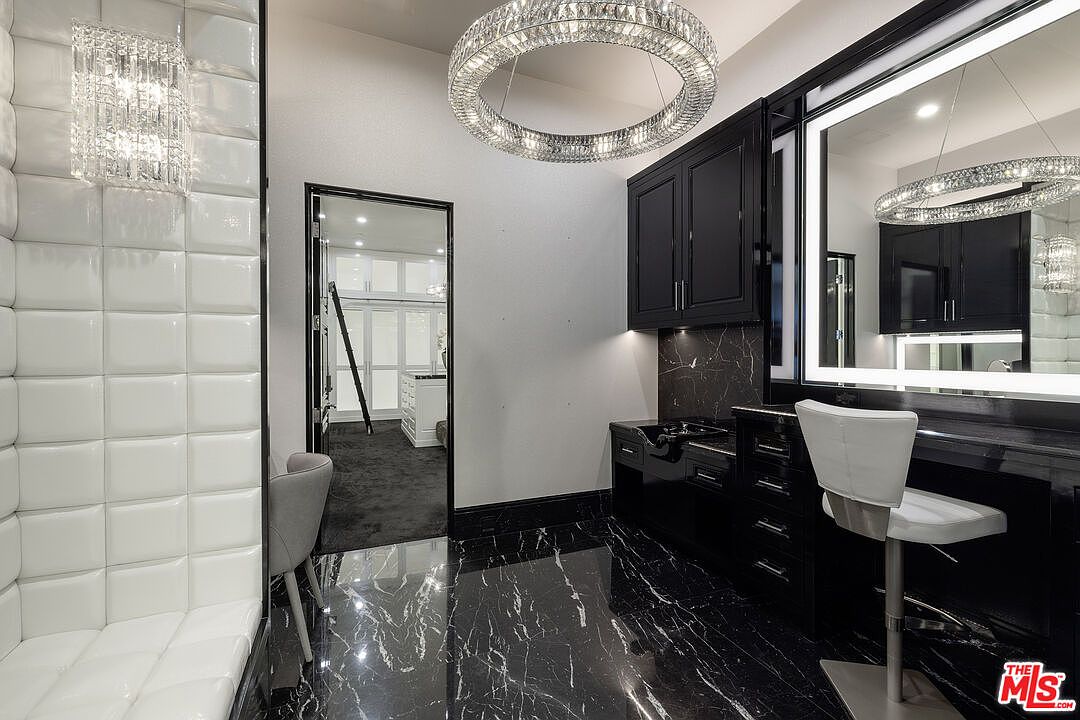 This luxurious primary bathroom features a high-contrast design with black cabinetry, dark stone flooring with white veining, and a white tufted wall panel. A sleek vanity area is equipped with a built-in sink and a large, illuminated mirror, complemented by a modern circular crystal chandelier. The space includes a doorway leading to a walk-in closet, creating a sophisticated and opulent atmosphere.