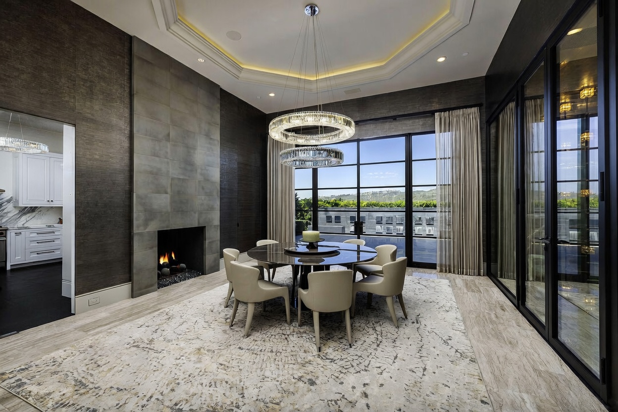 This elegant dining room features a circular table surrounded by six upholstered chairs, positioned beneath a dual-ring crystal chandelier. A prominent stone-clad chimney breast serves as a focal point, while large glass doors provide a view of the outdoor terrace and surrounding landscape. The space is defined by a textured wall treatment, light-toned stone flooring, and a large patterned area rug, creating a sophisticated and airy atmosphere.