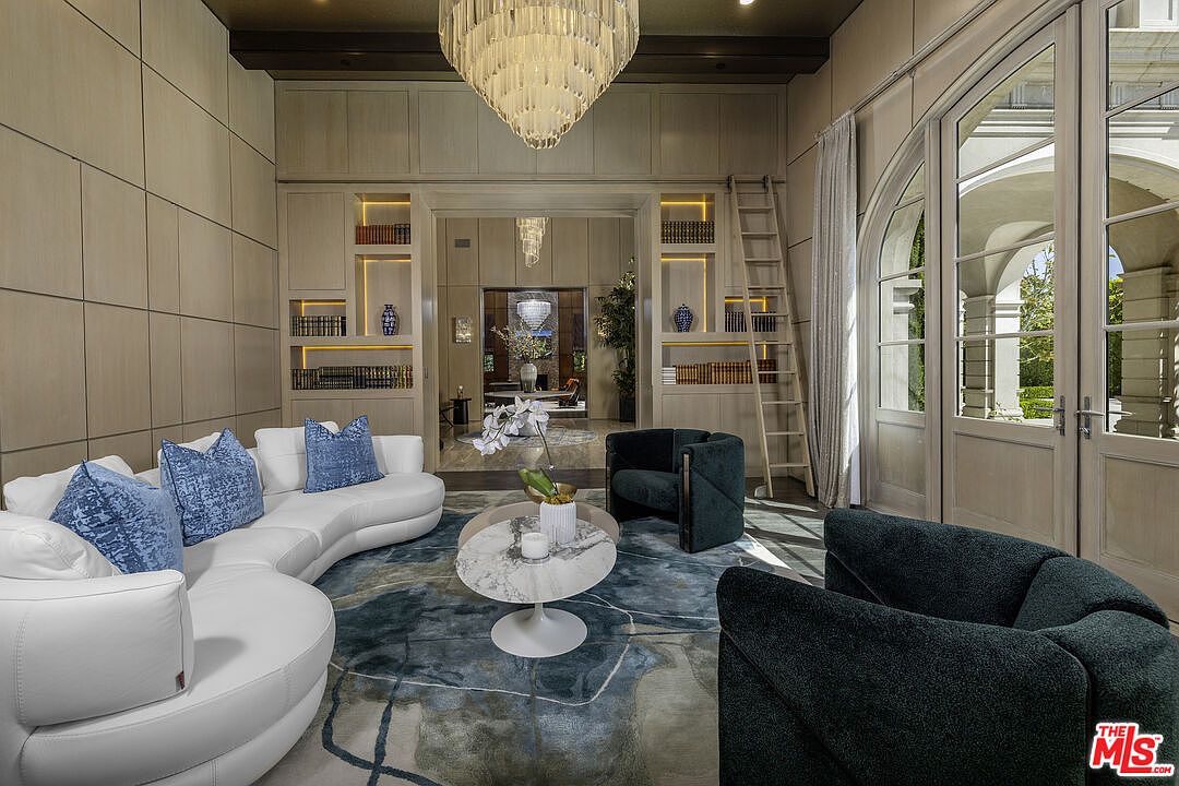 This sophisticated living room features a curved white sofa accented with blue patterned pillows, paired with dark charcoal armchairs and a circular stone-topped coffee table. The space is anchored by a large, abstract-patterned rug in shades of blue and grey, set against light wood-paneled walls with built-in shelving and a rolling library ladder. A grand crystal chandelier hangs from the ceiling, while tall, arched glass doors provide a view into an outdoor covered patio area.