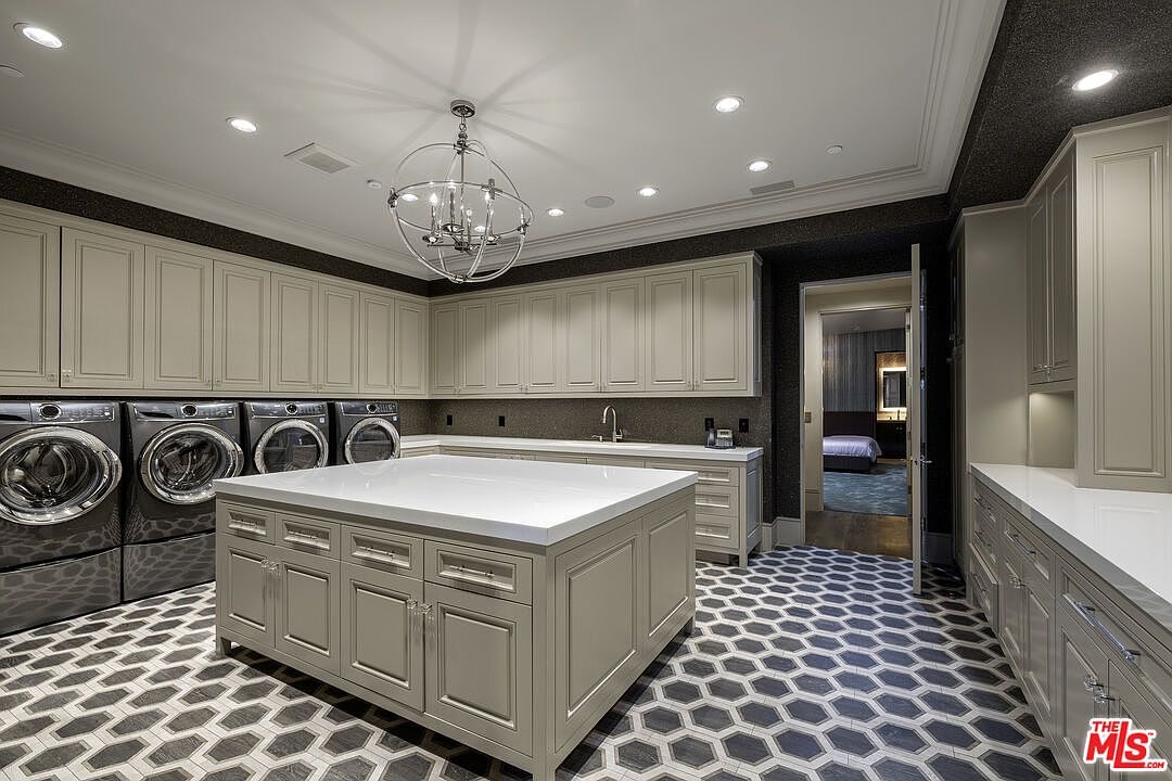 This spacious and well-appointed laundry room features a large central island with a white countertop, providing ample workspace for folding and organizing. The room is equipped with multiple high-end front-loading washers and dryers, extensive light-toned cabinetry for storage, and a striking geometric patterned tile floor. A decorative chandelier hangs above the island, and a doorway leads into an adjacent bedroom, creating a seamless flow between living spaces.