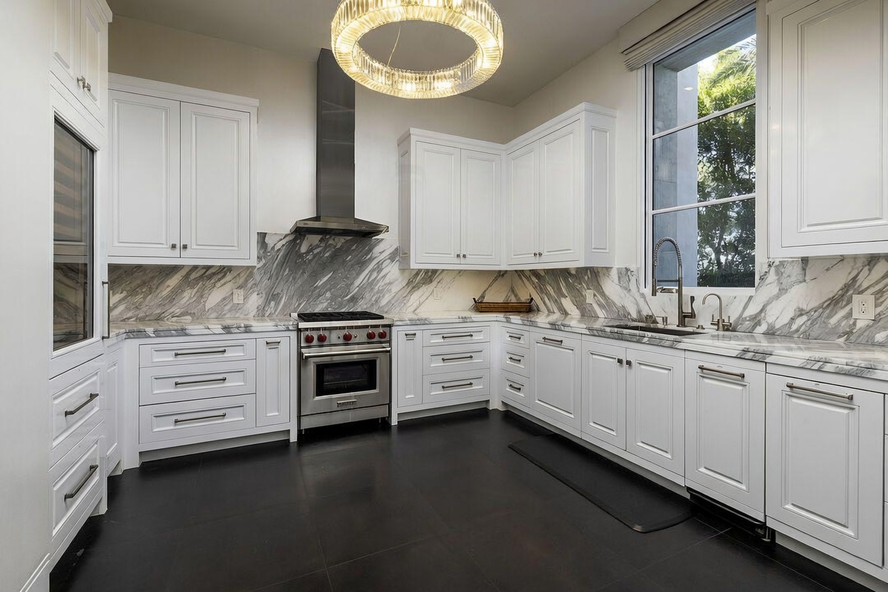 This bright, modern kitchen features crisp white cabinetry paired with striking, heavily veined stone countertops and backsplashes. The space is anchored by a professional-grade stainless steel range and a large, circular crystal chandelier that serves as a focal point above the center of the room. A tall window allows natural light to flood the area, highlighting the contrast between the white cabinetry and the dark, matte-finished floor tiles.