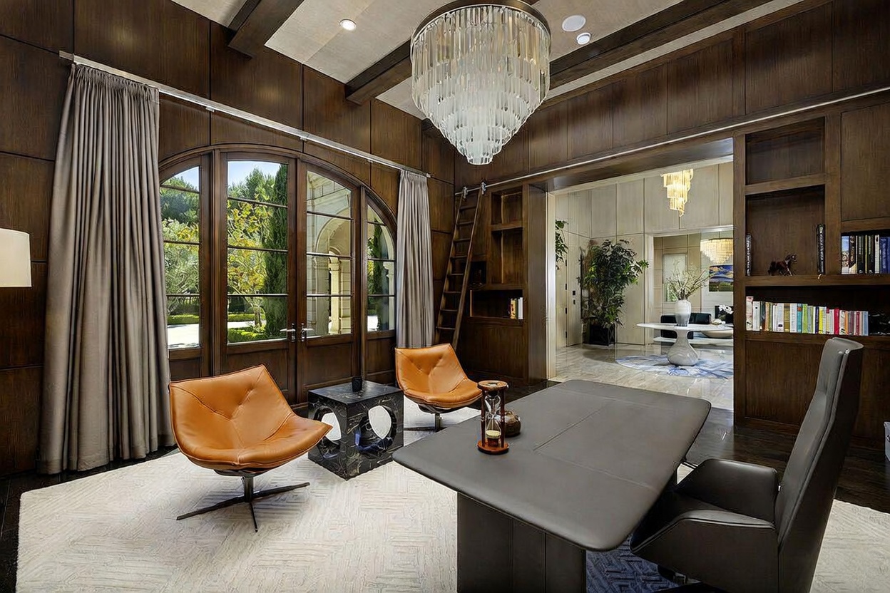 This sophisticated home office features rich, dark wood-paneled walls and a dramatic crystal chandelier suspended from a coffered ceiling. The room is furnished with a sleek, modern desk, two tan leather lounge chairs, and a geometric stone side table, all set upon a textured area rug. A rolling library ladder provides access to built-in shelving, while large arched glass doors offer a view into the lush greenery outside.