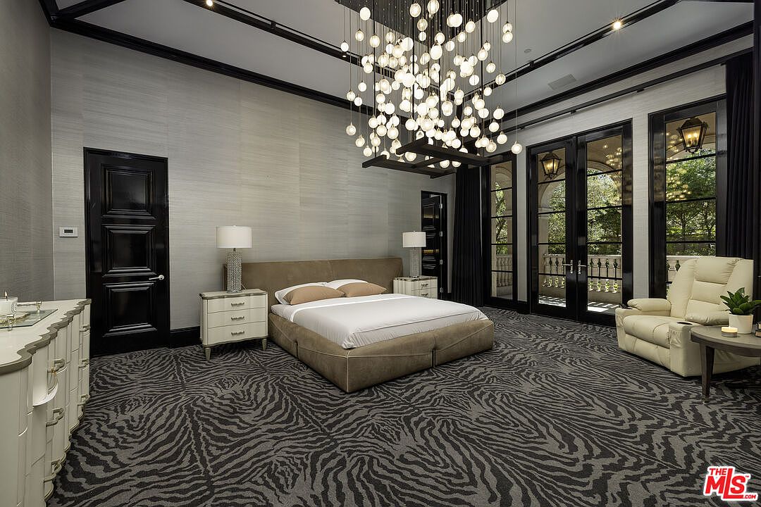 This spacious primary bedroom features a contemporary aesthetic with a bold, animal-print patterned carpet and a large, modern chandelier as the central focal point. The room includes a low-profile upholstered bed flanked by matching nightstands, a white dresser, and a comfortable leather armchair near the tall glass doors that open to an outdoor balcony. The walls are finished in a textured neutral wallpaper, complemented by dark trim and doors that provide a sophisticated contrast.