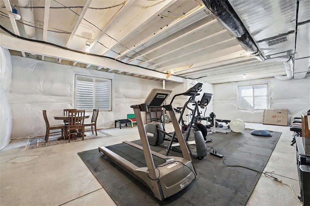 This spacious, unfinished basement serves as a versatile multi-purpose area, currently utilized as a home gym and casual dining space. The room features exposed ceiling joists, white-wrapped foundation walls, and a large black rubber mat covering the workout zone. The perspective is a wide-angle shot from the center of the room, capturing both the exercise equipment and the small dining set near the window.