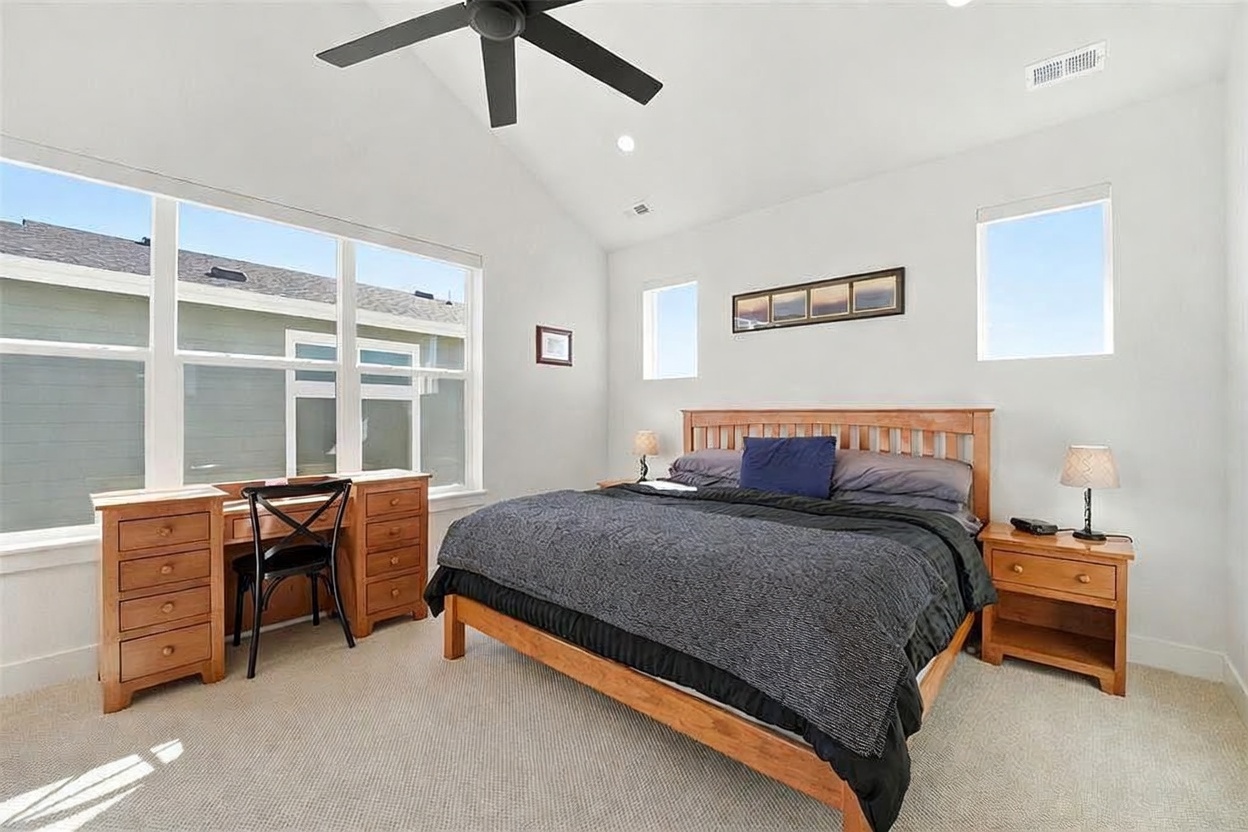This spacious primary bedroom features a vaulted ceiling with a central ceiling fan and neutral wall tones. The room is furnished with a wooden bed frame, matching nightstands, and a built-in desk area, all illuminated by natural light from large windows. The overall aesthetic is clean, functional, and inviting, creating a comfortable retreat.