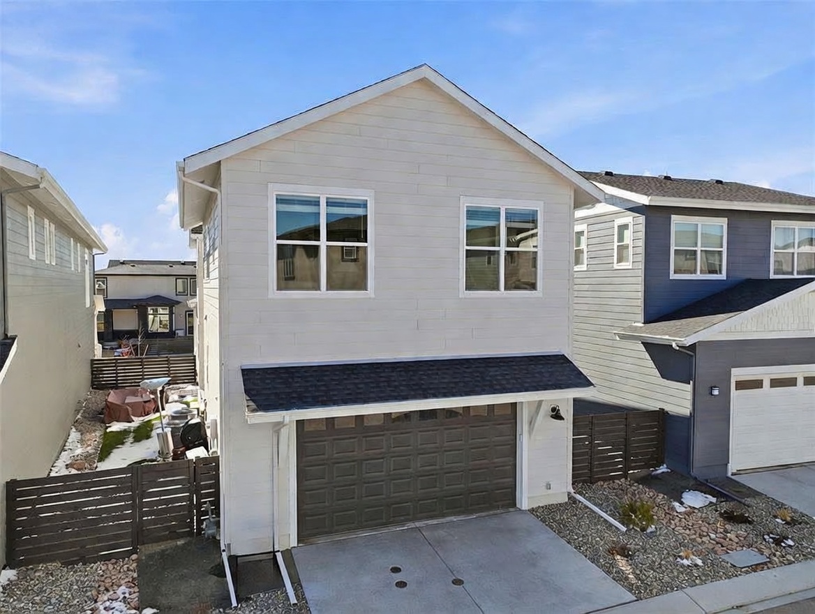 This is an elevated, angled exterior view of a two-story suburban home featuring light-colored horizontal siding and a dark-toned garage door. The property includes a small concrete driveway, a gravel landscaping bed, and a dark wooden privacy fence separating it from neighboring homes. The perspective captures the home's architectural profile against a clear blue sky, highlighting its compact, modern design.