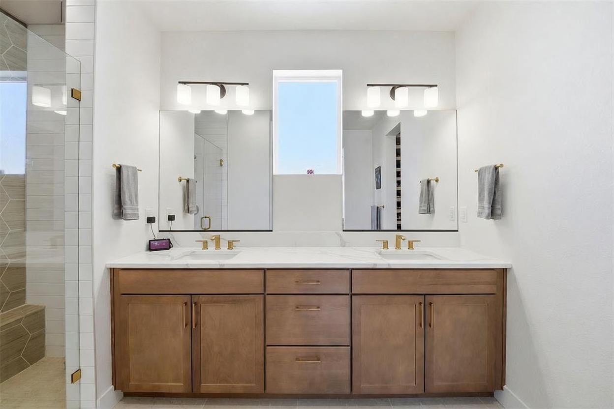 This primary bathroom features a double vanity with warm-toned wooden cabinetry, topped with a clean white countertop and dual undermount sinks. Two large, rectangular mirrors are mounted above the vanity, flanked by modern light fixtures, while a central window provides natural light. The space is finished with a neutral color palette, creating a bright, contemporary, and functional atmosphere.