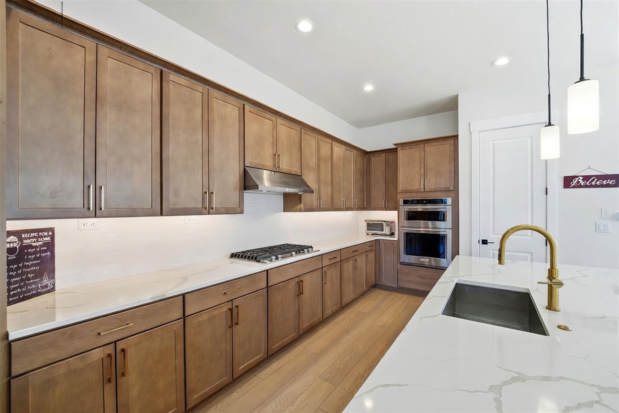 This modern kitchen features warm-toned wooden cabinetry paired with sleek white quartz countertops and a white subway tile backsplash. The space is equipped with stainless steel appliances, including a built-in double oven and a gas cooktop, and is illuminated by recessed lighting and elegant pendant lights over the island. The perspective captures the full length of the kitchen, highlighting the functional layout and the gold-toned faucet at the island sink.