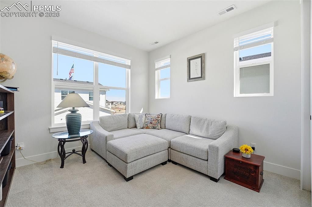 This bright and inviting living room features a large, light-gray sectional sofa with an ottoman, creating a cozy seating area. The space is illuminated by natural light from multiple windows, highlighting the neutral carpet and clean white walls. A small side table with a lamp and a rustic wooden chest add functional charm to this comfortable, well-lit room.
