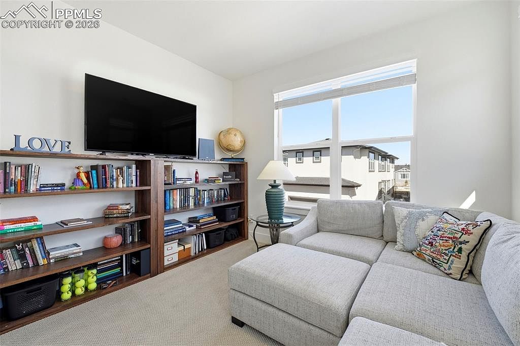 This cozy living room features a large, light-gray sectional sofa with a matching ottoman, positioned next to a bright window that offers a view of the neighborhood. A prominent wooden bookshelf unit spans the wall, housing a television, books, and decorative items like a globe. The space is well-lit and inviting, with a neutral color palette that creates a comfortable and functional atmosphere.