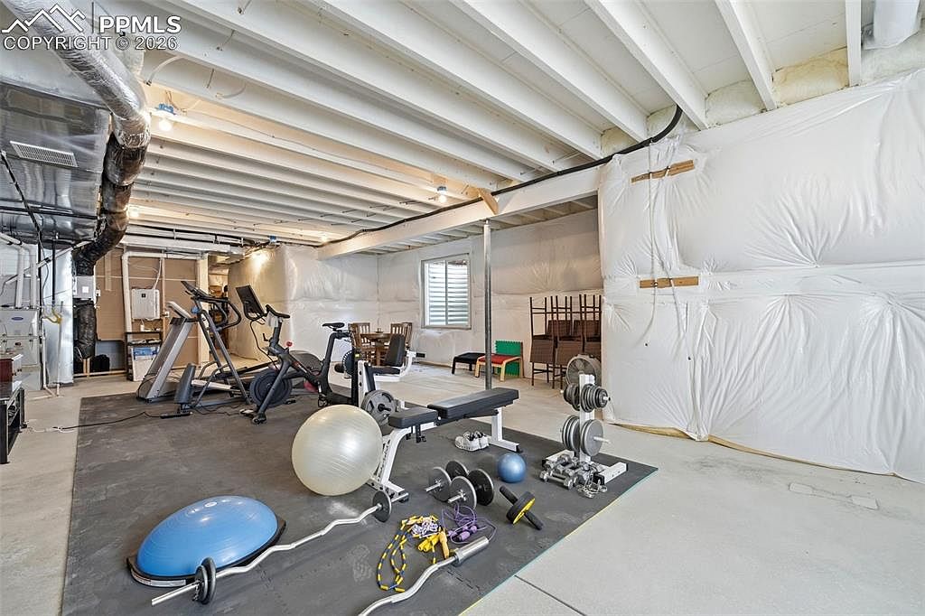 This spacious, unfinished basement has been converted into a functional home gym, featuring a large rubber mat area equipped with various exercise machines, free weights, and balance equipment. The room showcases an industrial aesthetic with exposed ceiling joists and white plastic-wrapped insulation on the walls, providing a raw, utilitarian feel. The perspective is a wide-angle shot from the floor level, capturing the entire workout zone and the transition into the unfinished storage area in the background.