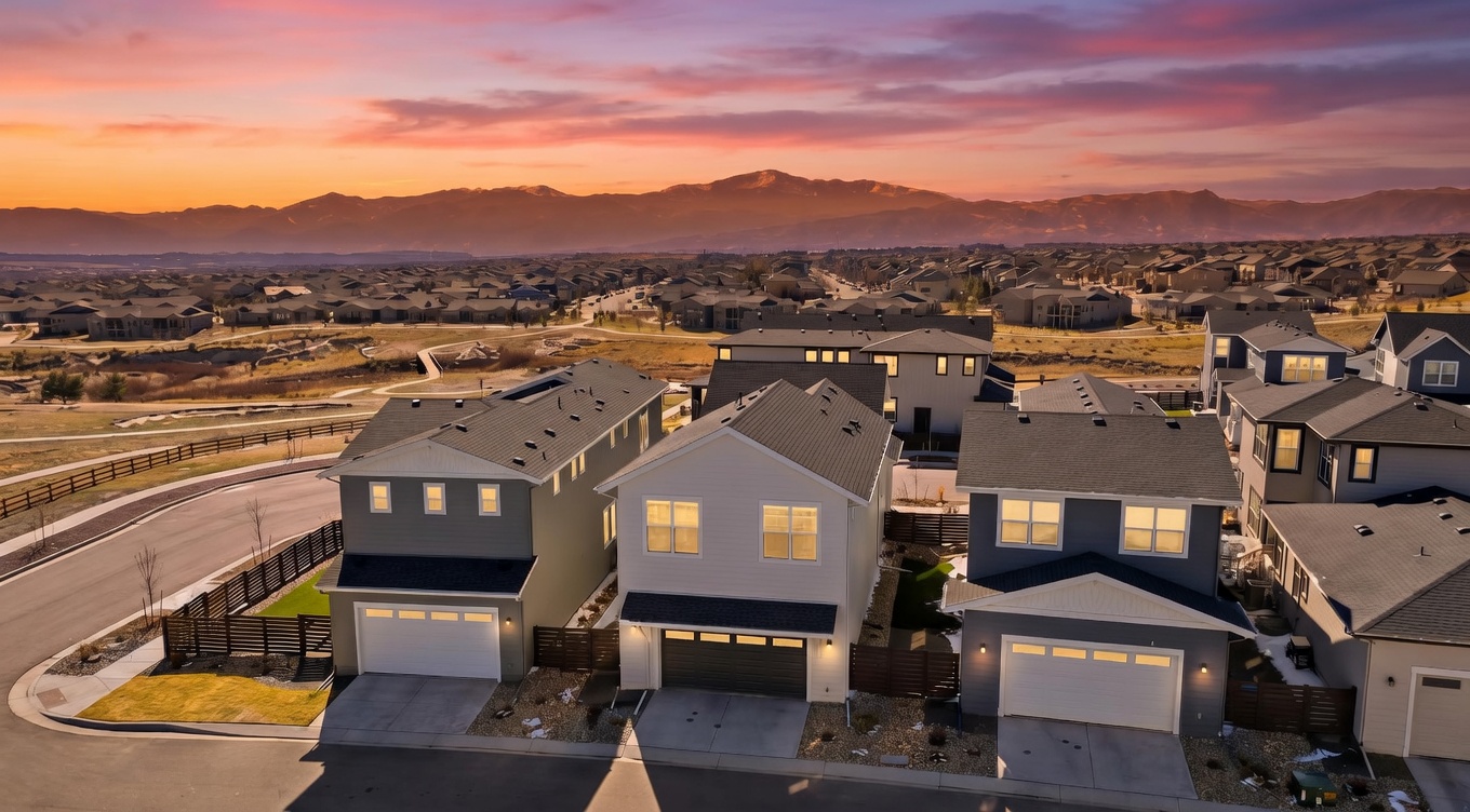 This aerial photograph captures a contemporary suburban neighborhood at sunset, frame-centered by three modern two-story homes with attached garages. The composition showcases the clean architectural lines of the houses, their front-facing garages, and the transition into a sprawling landscape with mountain views in the background. The warm, golden-hour sky enhances the appeal of this quiet residential community, highlighting the orderly street layout and pleasant suburban atmosphere.