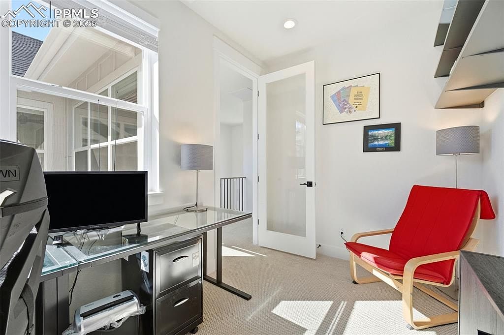 This bright and functional home office features a modern glass-top desk paired with a black filing cabinet and a vibrant red accent chair. The room is illuminated by natural light from a large window and a sleek ceiling light, creating an inviting workspace. A glass-paneled door leads to an adjacent hallway, while framed artwork adds a personal touch to the clean, neutral-toned walls.