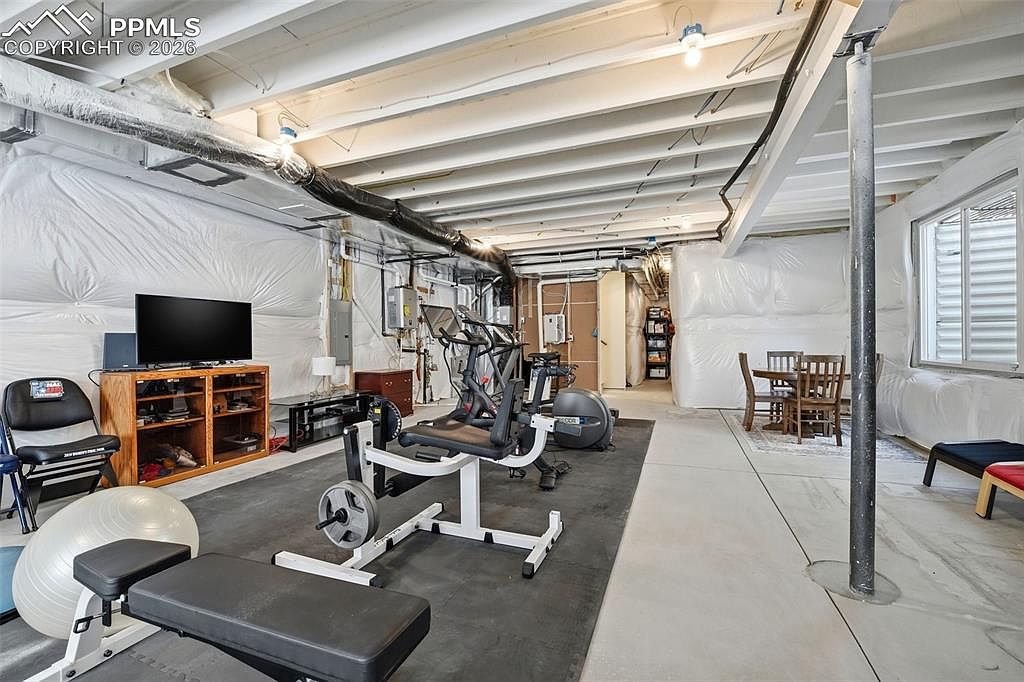 This spacious, unfinished basement has been converted into a functional home gym, featuring a variety of exercise equipment including a weight bench, stationary bike, and elliptical machine set on a rubber mat. The area also includes a small television setup and a dining table in the background, showcasing a versatile multi-purpose living space. The room features exposed ceiling joists, white plastic-wrapped walls, and a concrete floor, providing an industrial and utilitarian aesthetic.