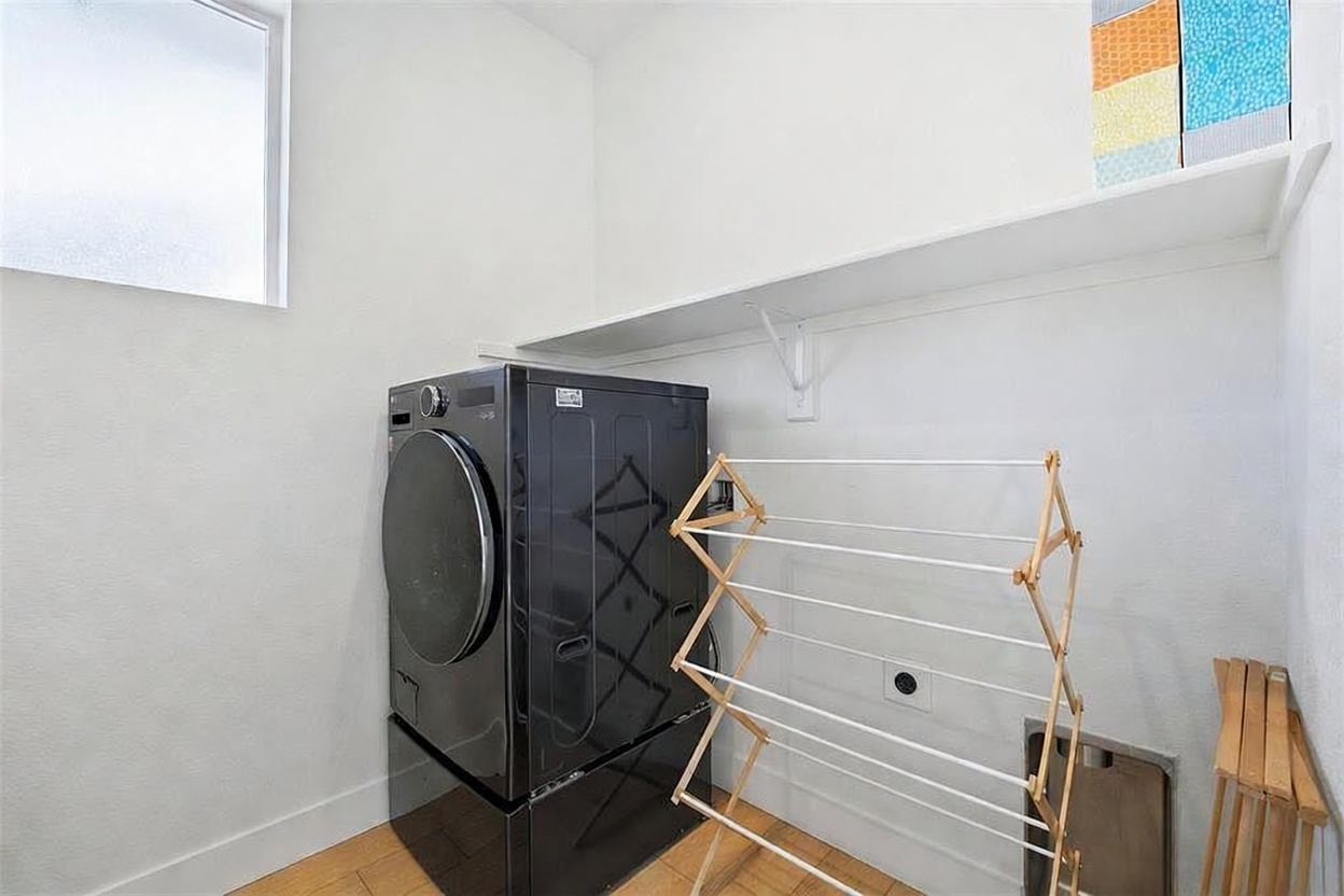 This functional laundry room features a sleek, black front-loading washer and dryer set positioned against a clean white wall. A wooden drying rack stands in the foreground, while a high shelf provides additional storage space above the appliances. The room is finished with light-toned wood flooring and a window that allows natural light to brighten the space.