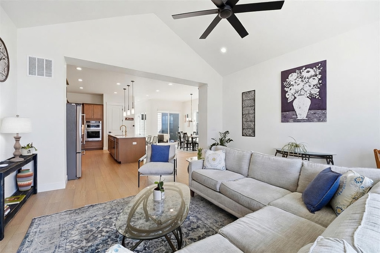 This bright and airy open-concept living room features a neutral color palette, a large sectional sofa, and a glass-topped coffee table set upon a patterned area rug. The space flows seamlessly into a modern kitchen with wood cabinetry and a central island, creating an inviting atmosphere for entertaining. Vaulted ceilings and a ceiling fan enhance the sense of spaciousness, while tasteful wall art adds a touch of personality to the clean, contemporary design.