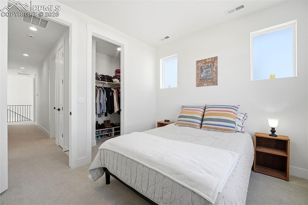 This bright and airy bedroom features a neutral color palette with a queen-sized bed adorned with striped accent pillows and a textured white quilt. To the left, an open doorway reveals a walk-in closet with organized shelving, while the room is illuminated by natural light from two windows and a bedside lamp. The perspective is from the foot of the bed, looking toward the closet and the hallway beyond, creating a clean and inviting atmosphere.