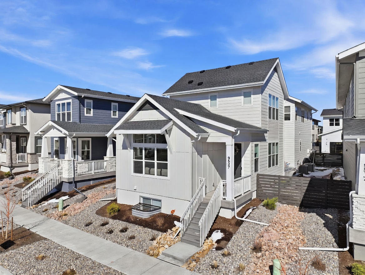 This image captures a modern, two-story residential home with a clean, white vertical siding exterior and a prominent front-facing gable. The property features a welcoming front porch with white railings, a small landscaped yard with gravel, and a clear view of the neighboring blue-sided home. The perspective is taken from a slightly elevated street-level angle, showcasing the home's contemporary architectural design and curb appeal under a bright, clear sky.