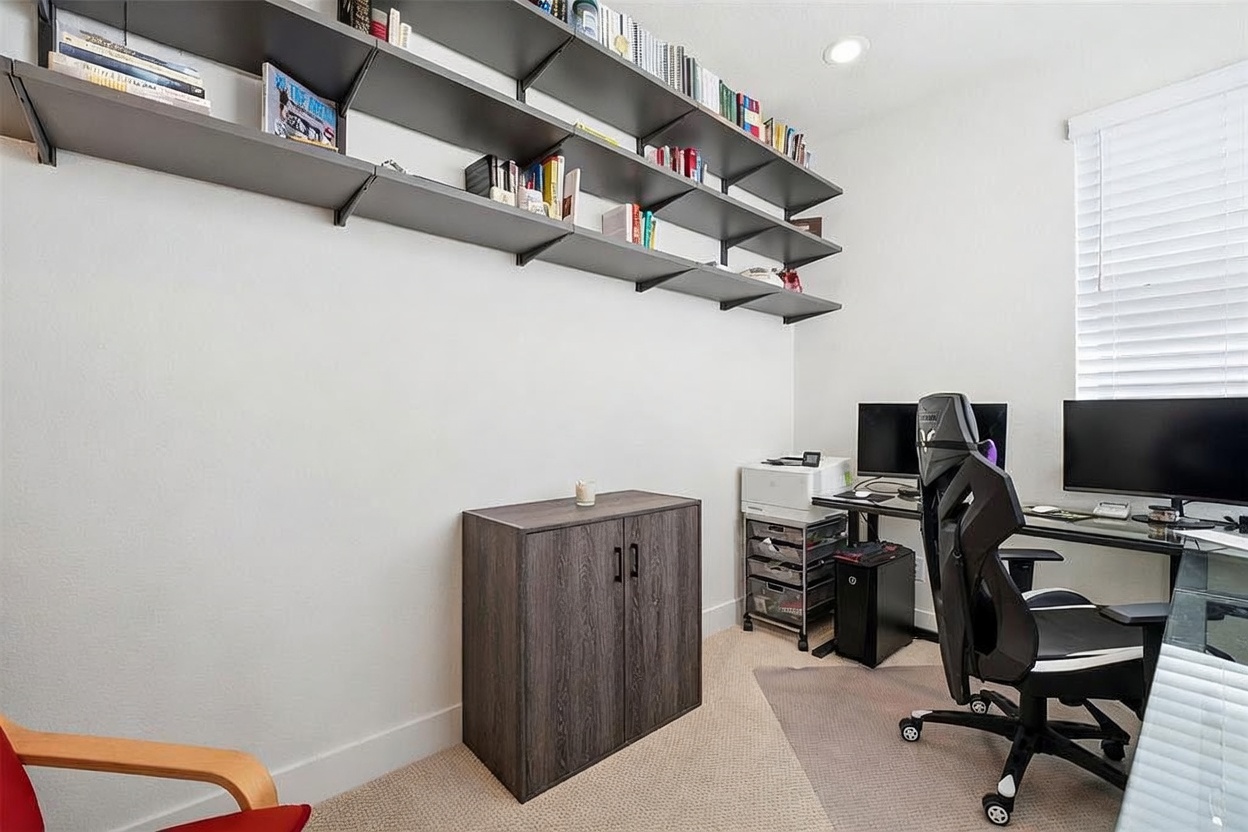 This functional home office features a clean, minimalist aesthetic with wall-mounted shelving units providing ample storage for books and decor. A dedicated workspace includes a modern desk setup with dual monitors and an ergonomic gaming chair, complemented by a separate storage cabinet and a printer station. The room is bright and organized, offering a productive environment with neutral tones and practical furniture placement.