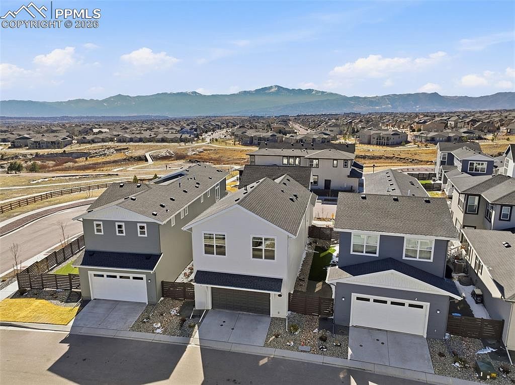 This high-angle aerial shot captures a row of modern, multi-story suburban homes in a newly developed neighborhood. The houses feature contemporary architectural designs with varied siding colors, attached garages, and small, fenced-in yards. In the background, a vast landscape of rolling hills and distant mountains provides a scenic and expansive horizon.