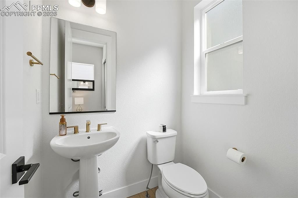 This bright and minimalist powder room features a clean white pedestal sink with gold-toned hardware and a matching faucet. A simple rectangular mirror hangs above the sink, reflecting the neutral-toned walls and a window that allows for natural light. The space is completed by a standard white toilet and a wall-mounted toilet paper holder, creating a functional and tidy guest bathroom.