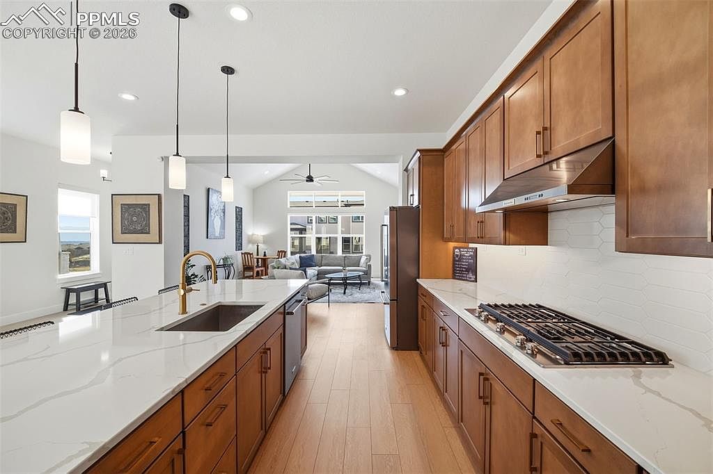 This modern, open-concept kitchen features warm wood cabinetry paired with sleek white quartz countertops and a white hexagonal tile backsplash. A large central island with a gold-toned faucet serves as a focal point, while pendant lighting adds a contemporary touch. The perspective looks from the kitchen toward the bright, airy living area, creating a seamless flow between spaces.