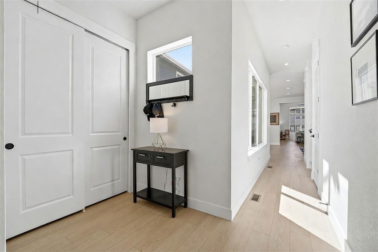 This bright and airy entryway leads into a long, clean-lined hallway featuring light-toned wood flooring and crisp white walls. To the left, a black console table sits beneath a wall-mounted mirror and coat rack, while a large closet with double doors provides ample storage. The perspective draws the eye down the hallway toward a distant living space, emphasizing the home's open and modern architectural flow.