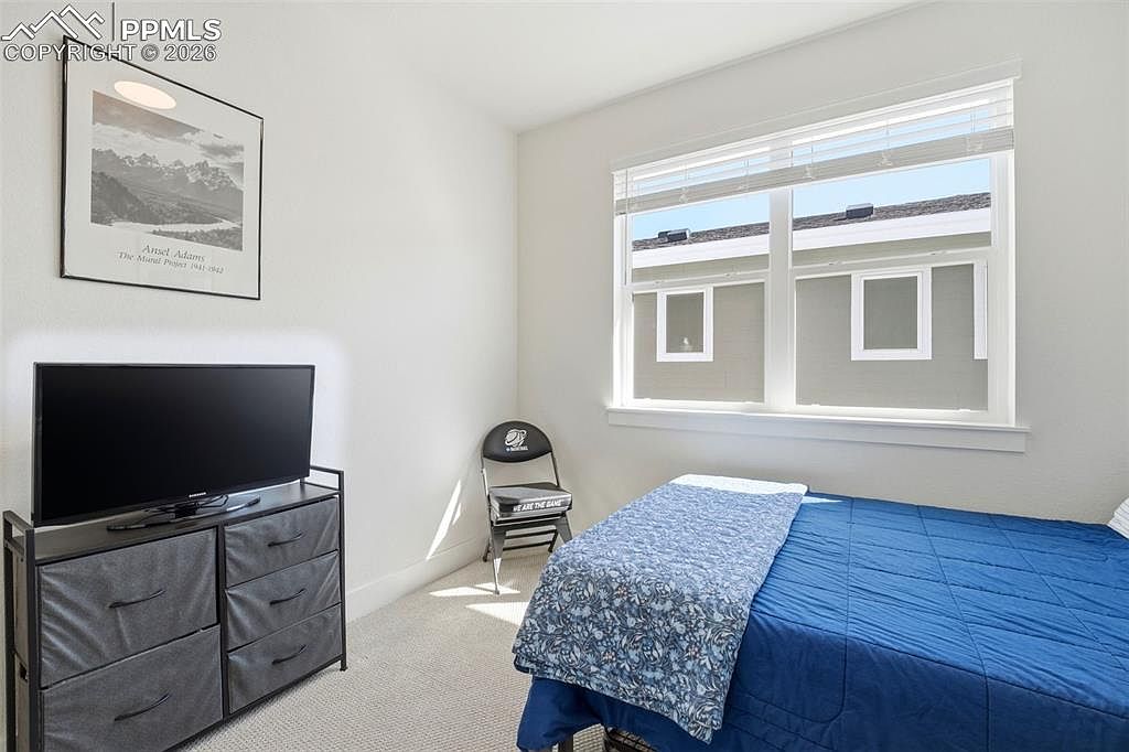 This bedroom features a simple, functional layout with a blue-quilted bed, a dark grey dresser topped with a flat-screen television, and a single folding chair in the corner. A large window allows natural light to brighten the room, while a framed Ansel Adams print adds a touch of classic art to the neutral-toned walls. The space is clean and minimalist, presenting a comfortable and straightforward guest room environment.
