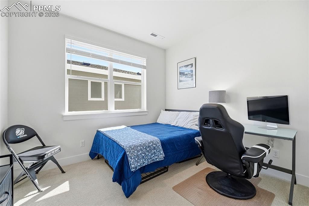 This compact bedroom features a twin-sized bed with a blue patterned comforter and a dedicated workspace with a gaming-style chair and desk. The room is brightly lit by a large window, creating a functional and clean space suitable for a guest room or home office. The neutral wall color and carpeted flooring provide a versatile foundation for various decor styles.