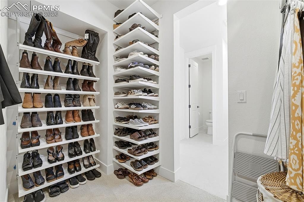 This organized walk-in closet features extensive white shelving units packed with a wide variety of footwear, including boots, sneakers, and sandals. The space is brightly lit and leads into a small, clean bathroom visible through the doorway in the background. A step stool and a patterned curtain add functional and decorative touches to this well-maintained storage area.