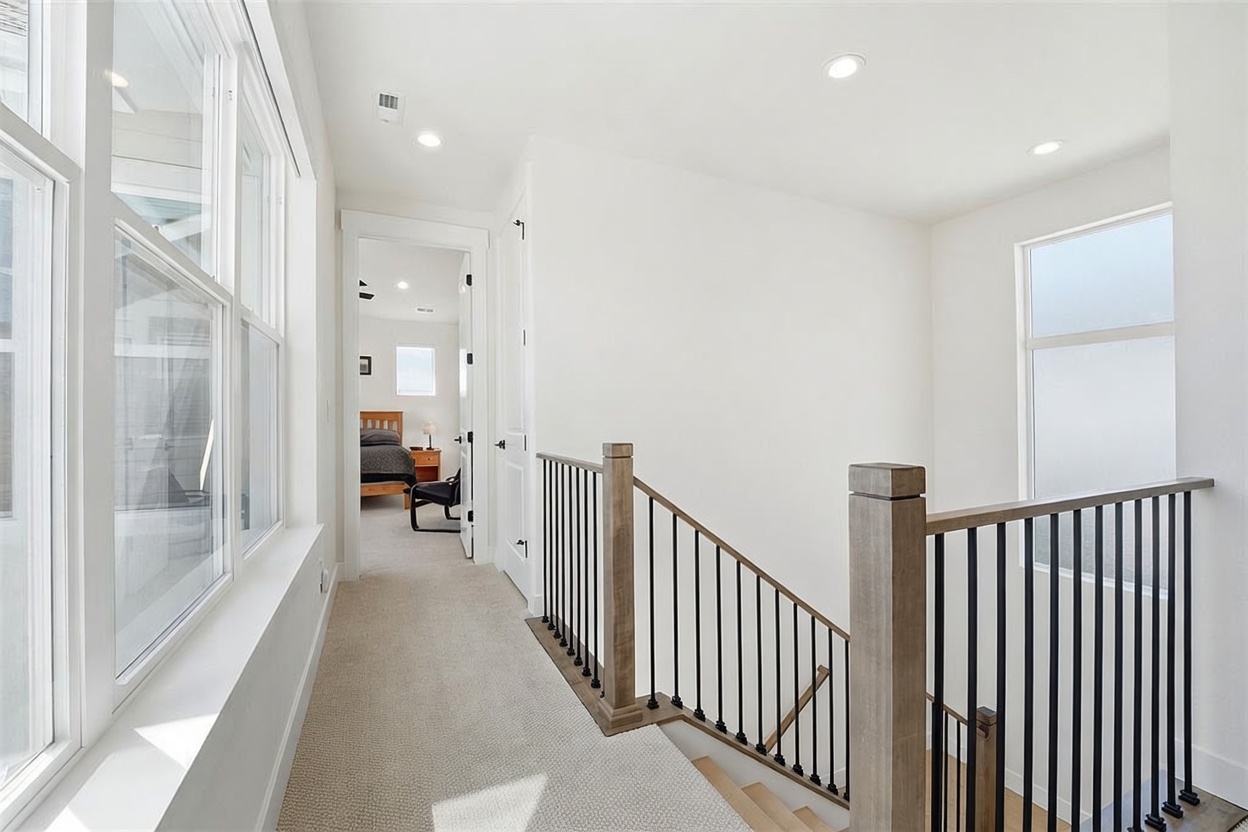 This bright and modern upstairs landing features a neutral color palette with light carpeting and crisp white walls. A wooden staircase with black metal balusters leads down, while a doorway opens into a bedroom, creating a clean and open transition space. The area is well-lit by recessed ceiling lights and large windows that allow for ample natural light.