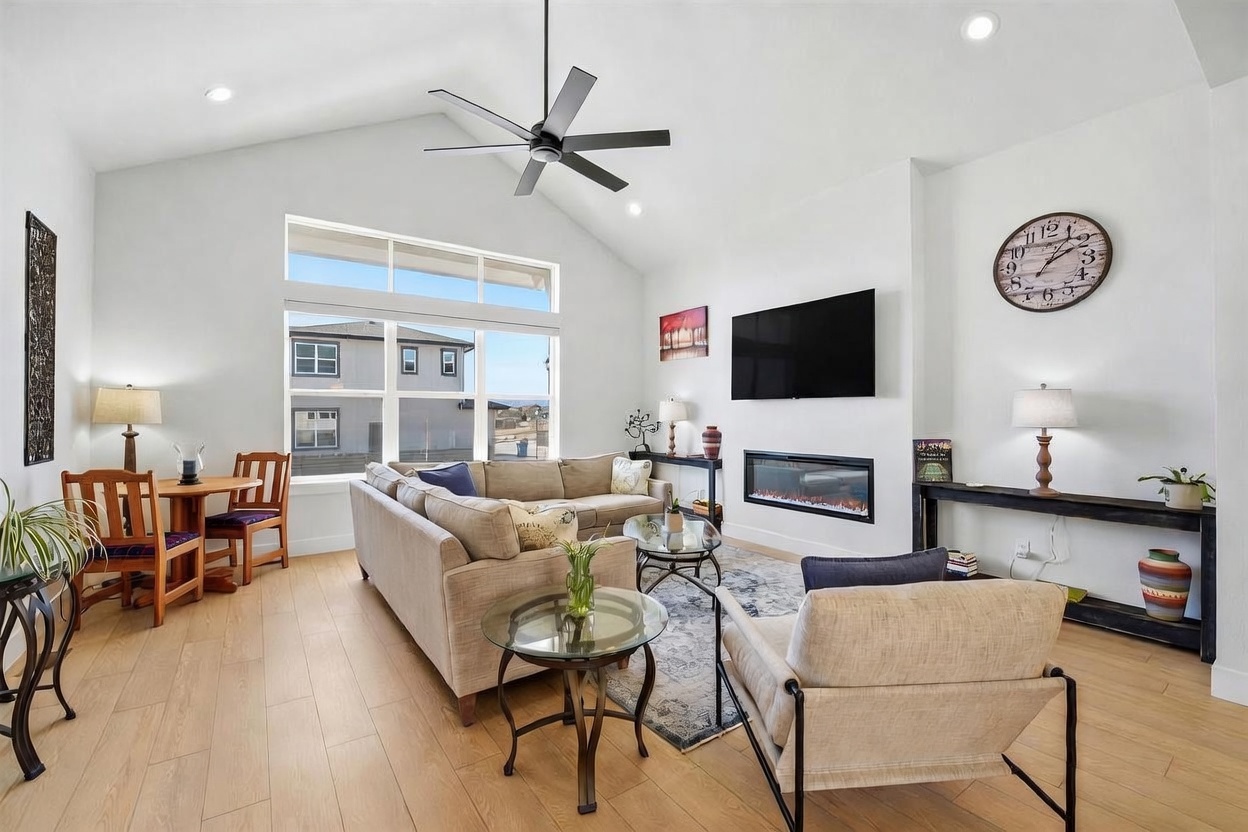 This bright and airy living room features a vaulted ceiling with a modern ceiling fan, light wood-look flooring, and a neutral color palette. The space is anchored by a comfortable sectional sofa, a glass-topped coffee table, and a sleek fireplace integrated into the wall beneath a mounted television. A dining area is tucked into the corner near large windows that provide ample natural light and a view of the neighboring homes.