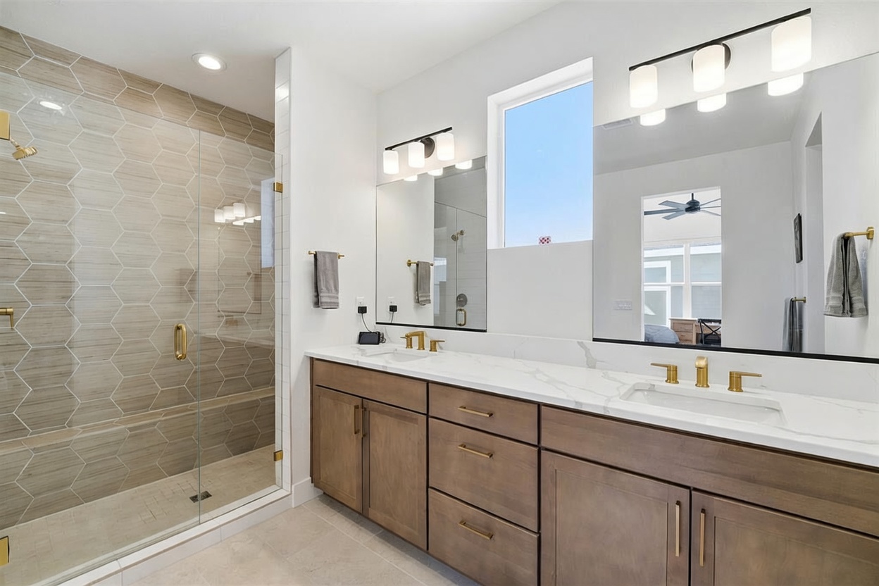 This modern primary bathroom features a double vanity with warm-toned wood cabinetry, gold hardware, and a white marble countertop. To the left, a spacious walk-in shower is enclosed by glass and tiled with elegant, light-colored hexagonal patterns. The room is bright and airy, enhanced by large mirrors, contemporary light fixtures, and a clean, minimalist aesthetic.