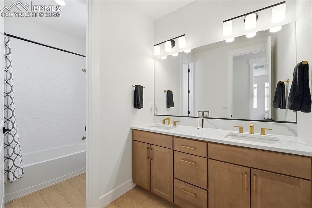 This modern primary bathroom features a double vanity with light wood cabinetry, a white quartz countertop, and gold-toned fixtures. A large, frameless mirror spans the wall above the sinks, complemented by contemporary light fixtures, while a separate shower area with a patterned curtain is visible to the left. The space is bright and clean, showcasing a neutral color palette and high-quality finishes.
