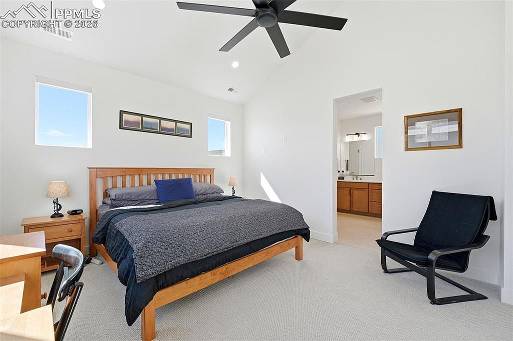 This bright and airy primary bedroom features a wooden bed frame with a dark blue and grey bedding set, complemented by a modern black accent chair. The room is illuminated by a ceiling fan and natural light from two windows, with an open doorway leading into an attached bathroom. The space has a clean, minimalist aesthetic with neutral walls and carpeted flooring.
